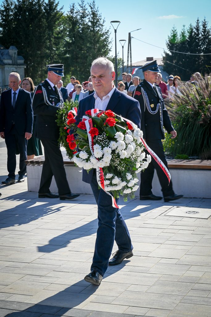 Mężczyzna w granatowej marynarce niesie biało-czerwoną wiązankę kwiatów, za nim ludzie, w tym dwaj mężczyźni w mundurach, na tle zielonych drzew i jasnego nieba.