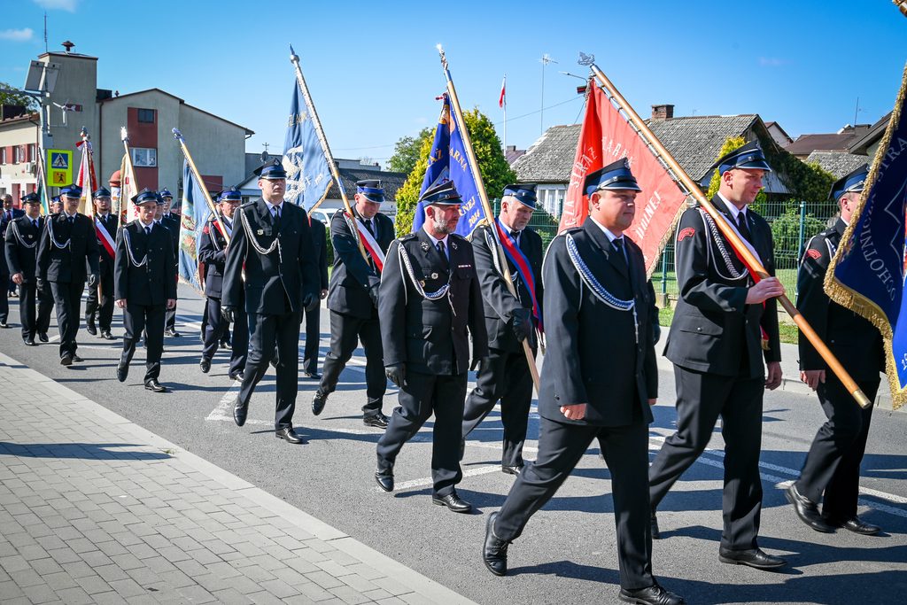Mężczyźni w czarnych mundurach i czapkach z daszkiem niosą kolorowe flagi, maszerują po ulicy w słoneczny dzień, w tle domy i błękitne niebo.