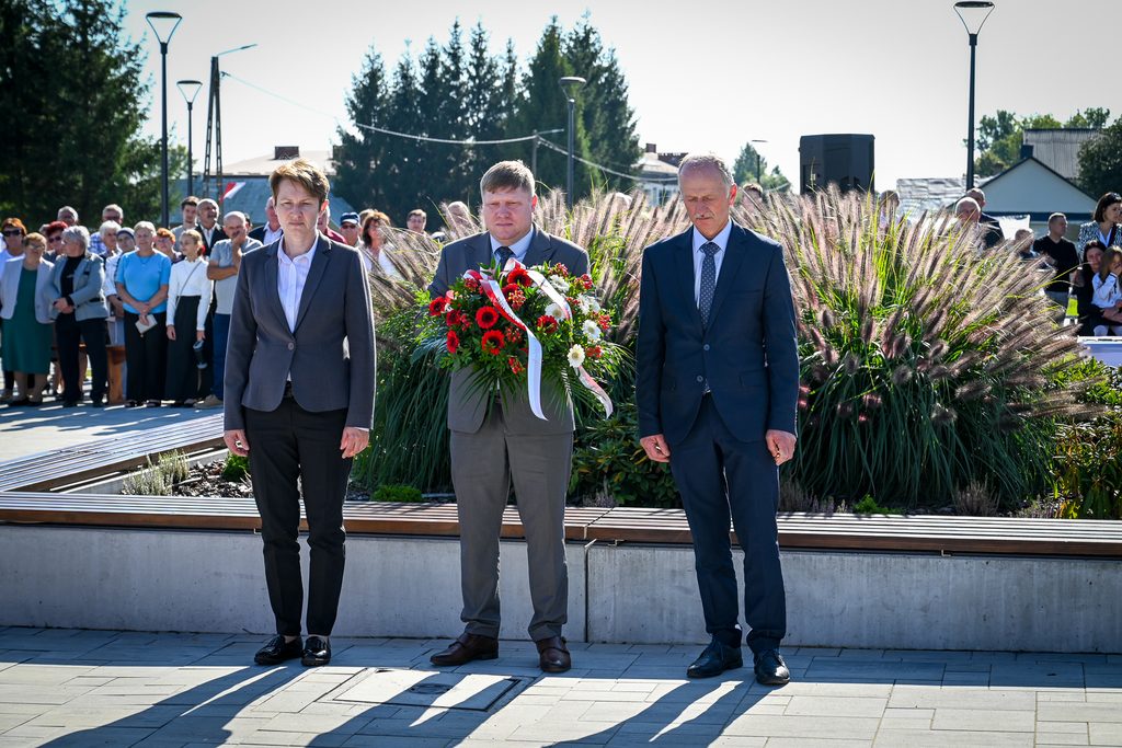 Trzy osoby w garniturach stojące na tle wysokiej trawy ozdobnej, mężczyzna pośrodku trzyma bukiet czerwonych i białych kwiatów, w tle grupa ludzi i latarnie uliczne.