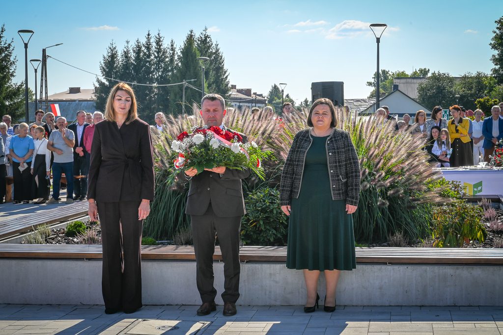 Trzy osoby stoją na tle zielonych traw ozdobnych, mężczyzna w środku trzyma bukiet czerwonych i białych kwiatów, za nimi stoi grupa ludzi, niebo jasne z nielicznymi chmurami.