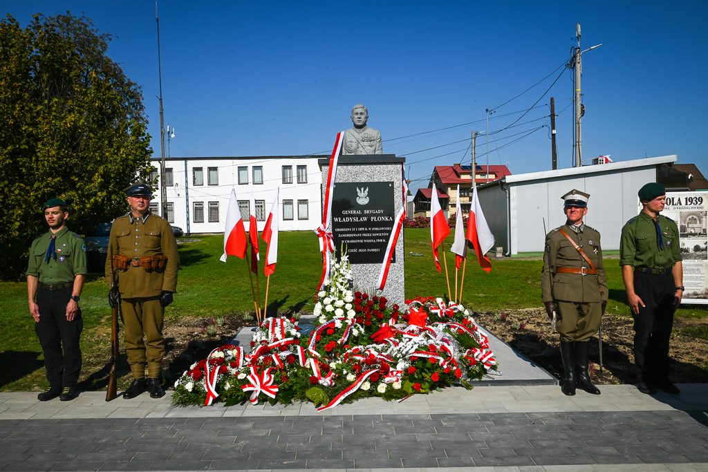 Pomnik generała Władysława Płonki z biało-czerwonymi flagami i kwiatami, wokół stoją cztery osoby w mundurach wojskowych, zielona trawa, niebieskie niebo.
