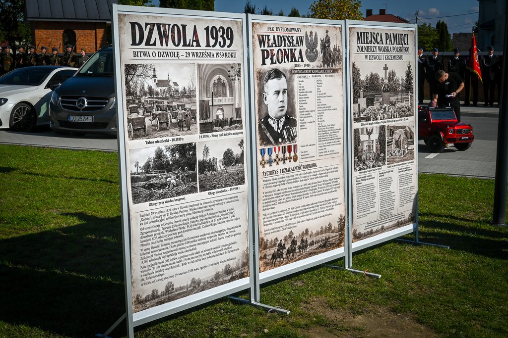 Trzy tablice informacyjne na trawie, z historycznymi zdjęciami i tekstem, w tle ludzie w mundurach i samochody.