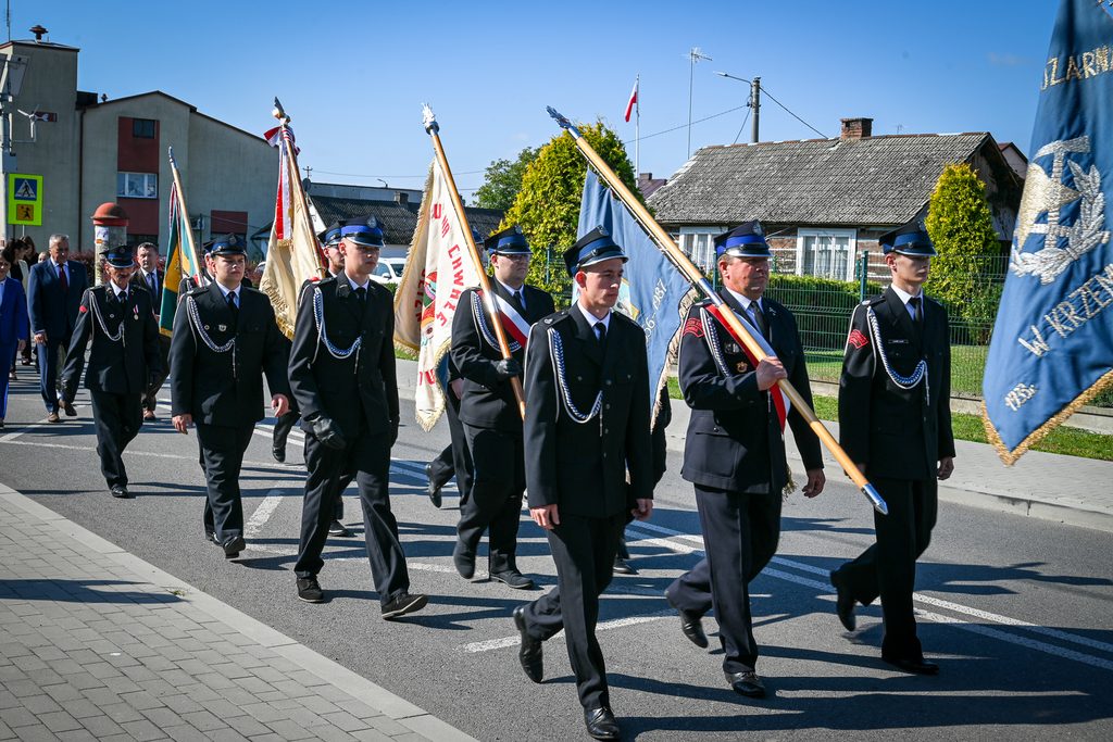 Grupa mężczyzn w mundurach strażackich idąca w pochodzie, niebieskie niebo, budynki mieszkalne i chodnik z kostki brukowej.