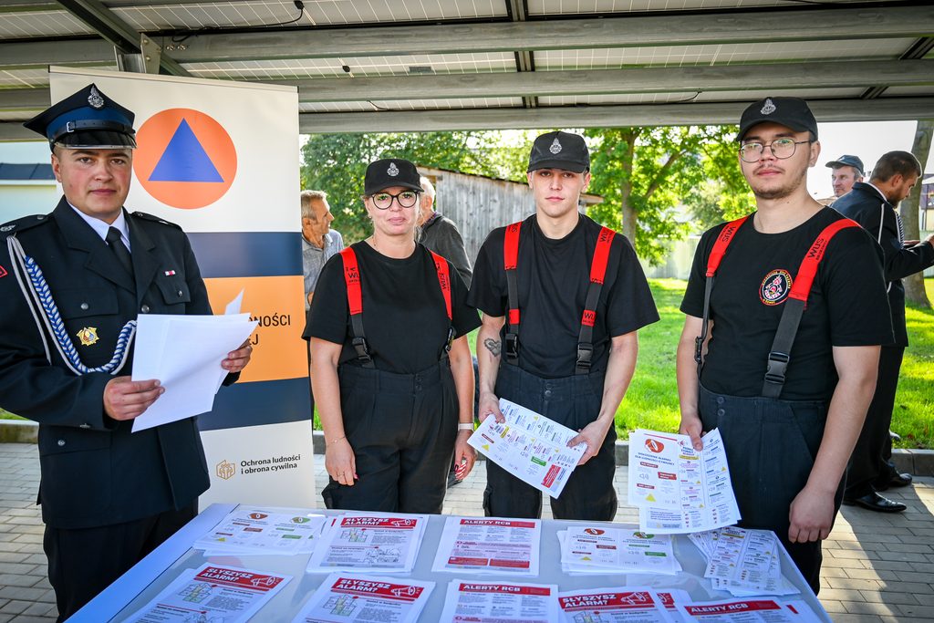 Cztery osoby w mundurach i czapkach stoją przy stole z ulotkami informacyjnymi, za nimi baner z symbolem ochrony ludności, w tle drzewa i drewniana szopa.