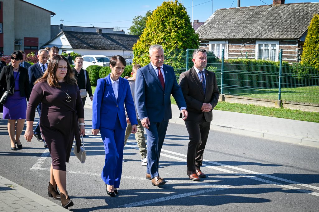 Grupa ludzi w eleganckich strojach idąca ulicą, w tle domy, zieleń i niebieskie niebo.