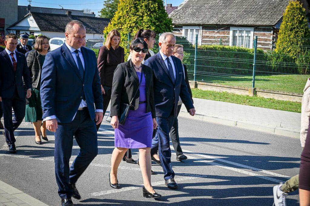 Grupa ludzi w eleganckich strojach idąca chodnikiem i ulicą w słoneczny dzień, za nimi domy, zieleń i metalowe ogrodzenie.