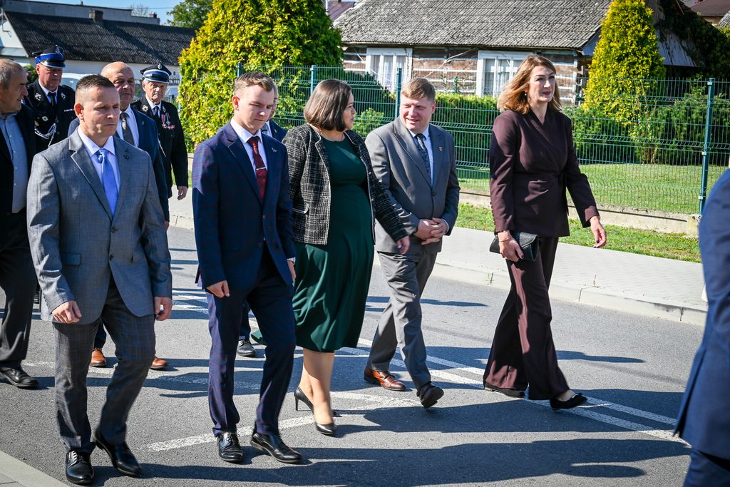 Grupa ludzi w eleganckich ubraniach idąca ulicą, w tle zieleń i drewniany dom z płotem metalowym.
