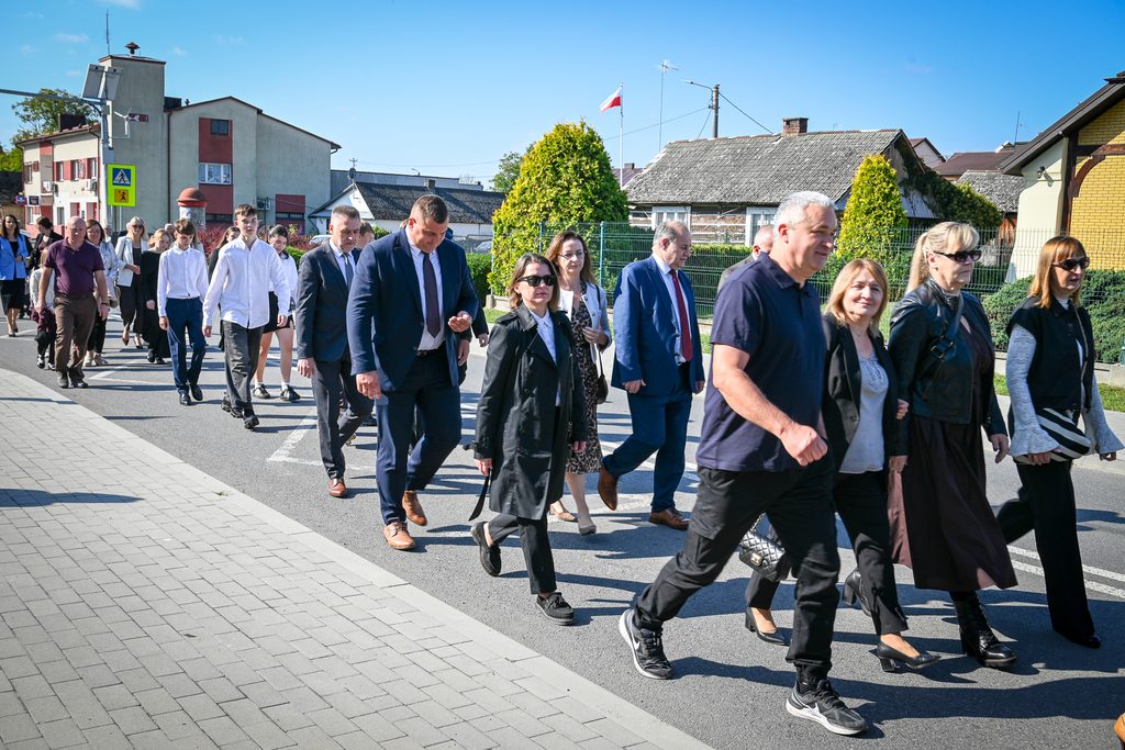 Grupa osób w różnym wieku w ubraniach formalnych i codziennych idzie chodnikiem i jezdnią przy słonecznej pogodzie, w tle zabudowania mieszkalne i polska flaga.