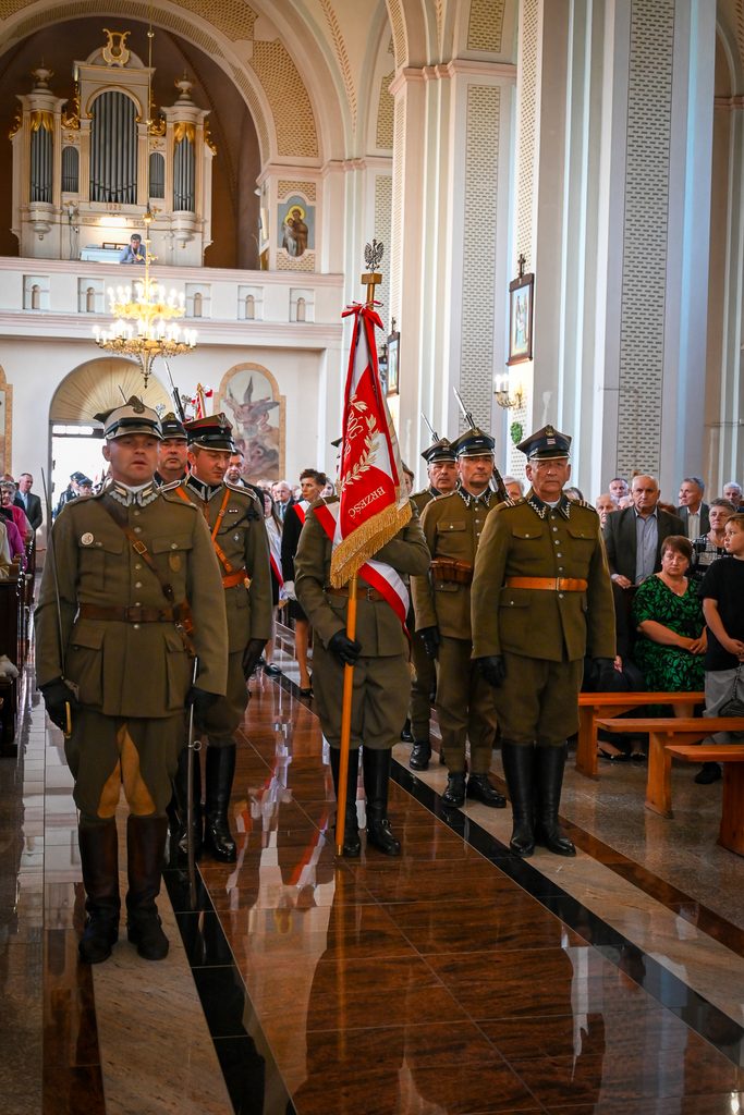 Grupa mężczyzn w historycznych mundurach wojskowych maszerujących w jasnym wnętrzu kościoła z czerwoną flagą, w tle ludzie siedzący na ławkach i organy.