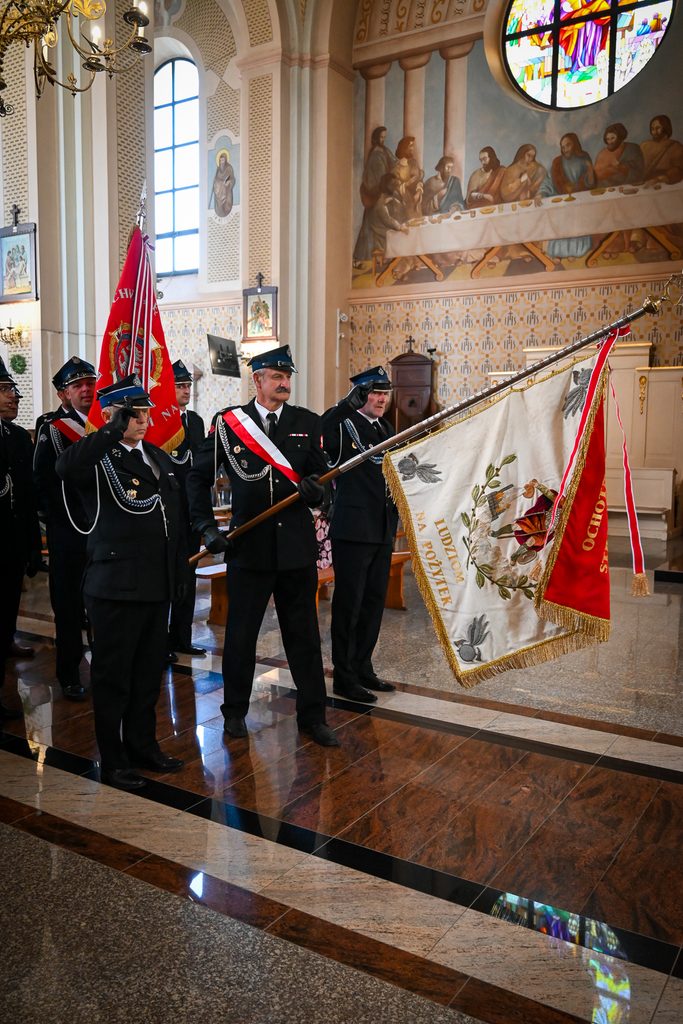 Grupa mężczyzn w mundurach strażackich w kościele, trzymających czerwono-biały sztandar, z malowidłem Ostatniej Wieczerzy i witrażem w tle.