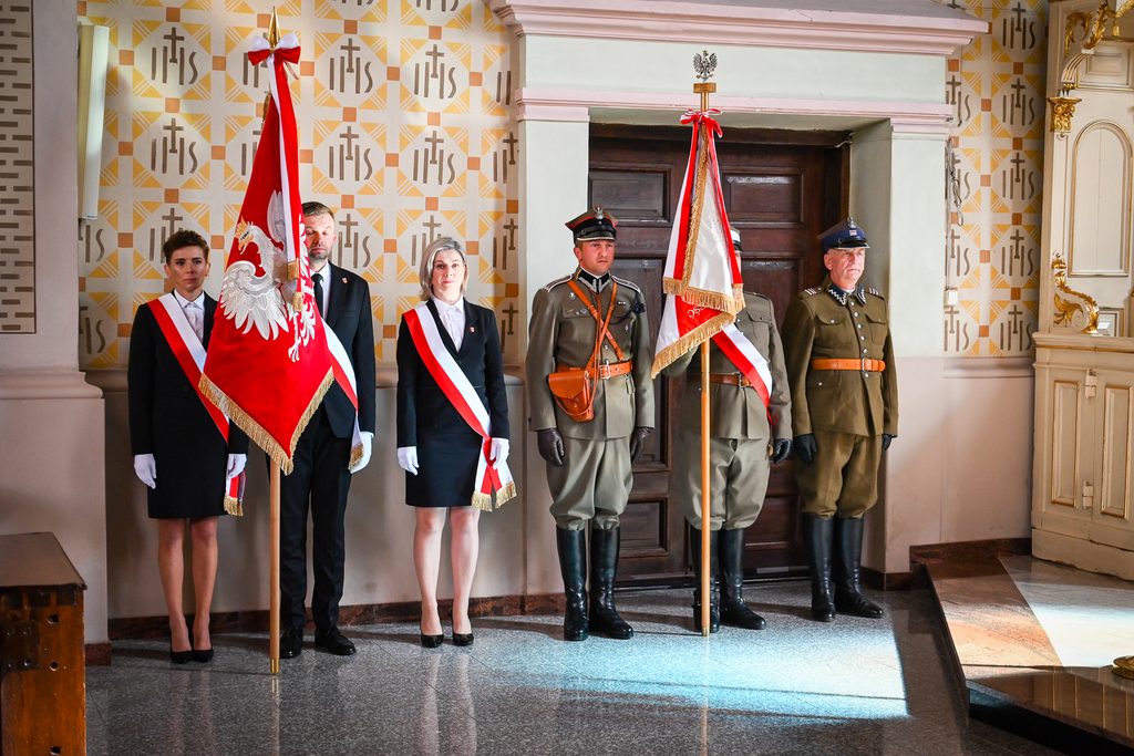 Grupa sześciu osób w mundurach i galowych strojach, trzy z nich trzymają flagi, na tle jasnej ściany z wzorem i ciemnych drzwi.