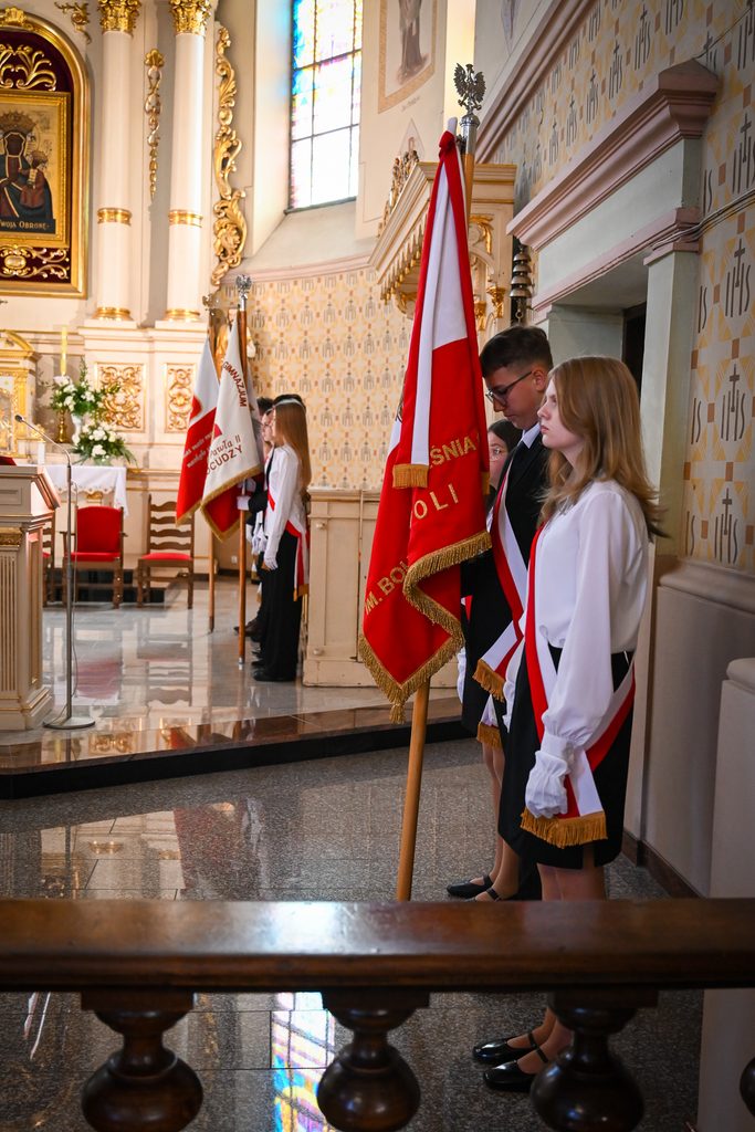 Młodzież w białych bluzkach i czarnych spódnicach oraz garniturach z biało-czerwonymi szarfami trzymająca czerwone i białe sztandary w ozdobionym złotem wnętrzu kościoła.