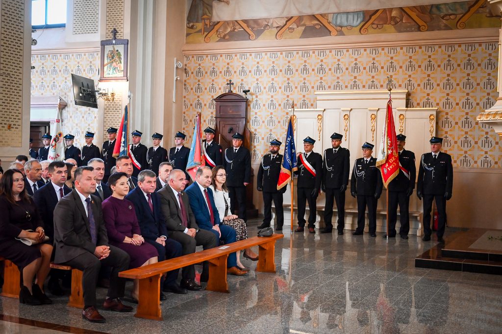Mężczyźni w mundurach strażackich z flagami i siedzący ludzie w formalnych strojach na ławce w jasnym wnętrzu z dekoracyjnymi ścianami i marmurową podłogą.