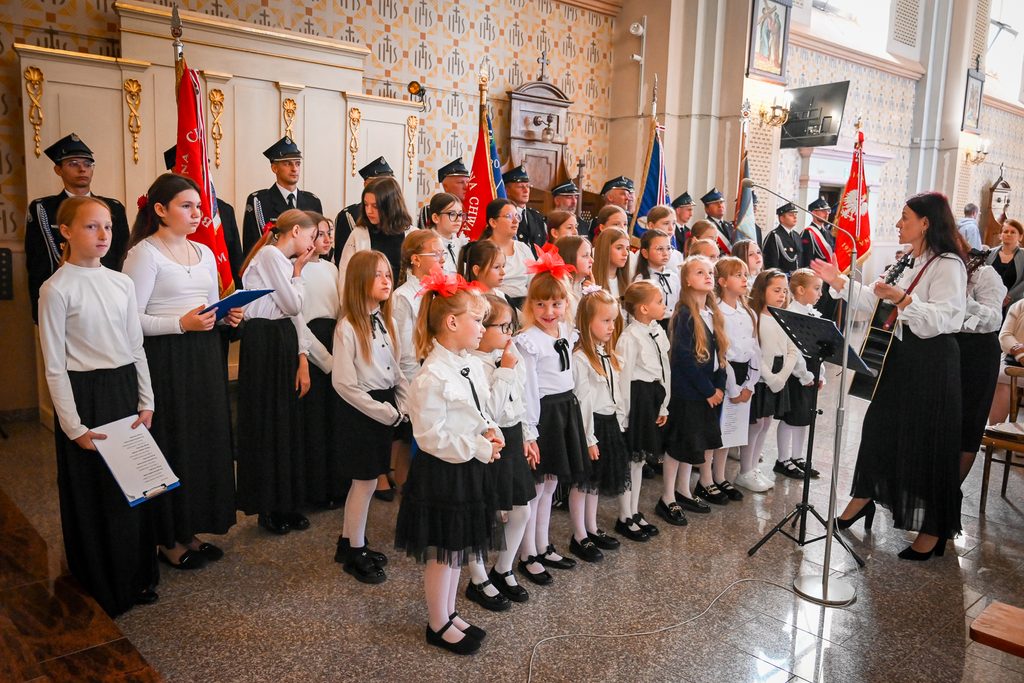 Grupa dzieci w białych bluzkach i czarnych spódnicach śpiewa, kobieta z gitarą dyryguje, w tle mężczyźni w mundurach z flagami w jasnym wnętrzu.