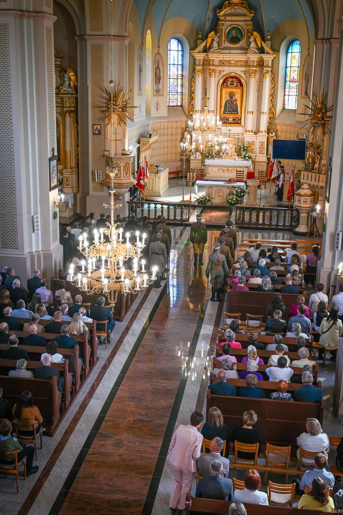 Wnętrze jasnego kościoła z bogato zdobionym ołtarzem, dużymi żyrandolami, wypełnione ławkami z siedzącymi ludźmi, żołnierze idący w kierunku ołtarza.