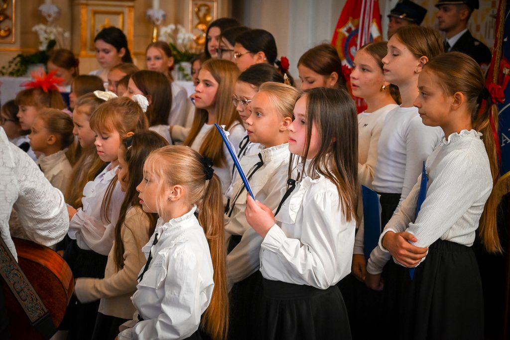 Grupa dziewczynek w białych bluzkach i czarnych spódnicach, niektóre trzymają niebieskie teczki, w tle fragment dekoracji i mężczyźni w mundurach.