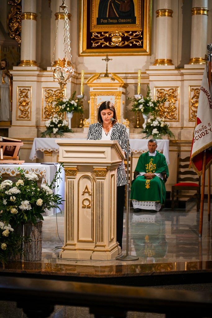 Kobieta w czarno-białej marynarce przy ambonie z symbolami Alfa i Omega, w tle kapłan w zielonym ornacie, kwiaty, złote zdobienia i obraz religijny.