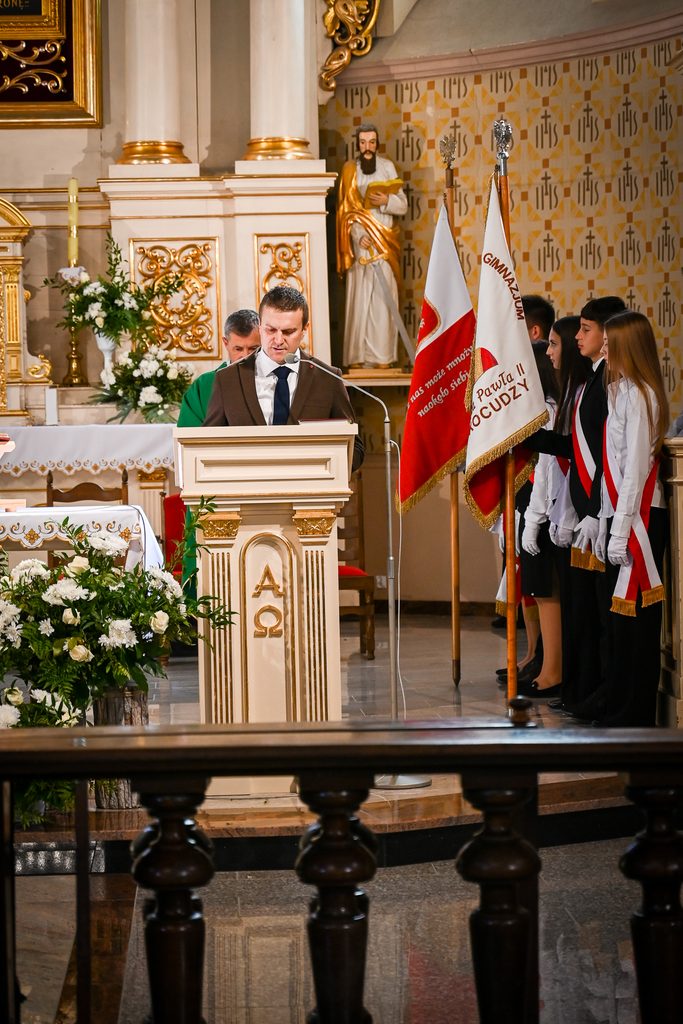 Mężczyzna w brązowej marynarce przy ambonie w jasnym wnętrzu kościoła, obok grupa młodzieży w białych rękawiczkach z biało-czerwonymi szarfami, flagi, kwiaty, złote zdobienia.