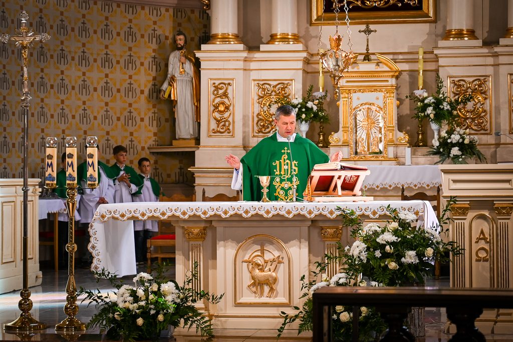 Kapłan w zielonej szacie za ołtarzem z białą koronkową serwetą, złote zdobienia, srebrny kielich, świece, białe kwiaty, w tle chłopcy w białych albach z zielonymi pelerynami.
