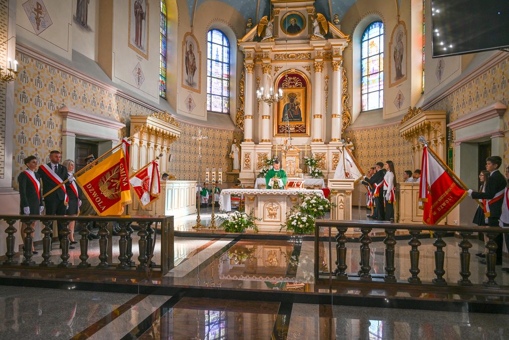 Wnętrze kościoła z ołtarzem, kapłan w zielonych szatach, osoby z flagami po obu stronach, witraże, złote zdobienia, białe kwiaty na ołtarzu.