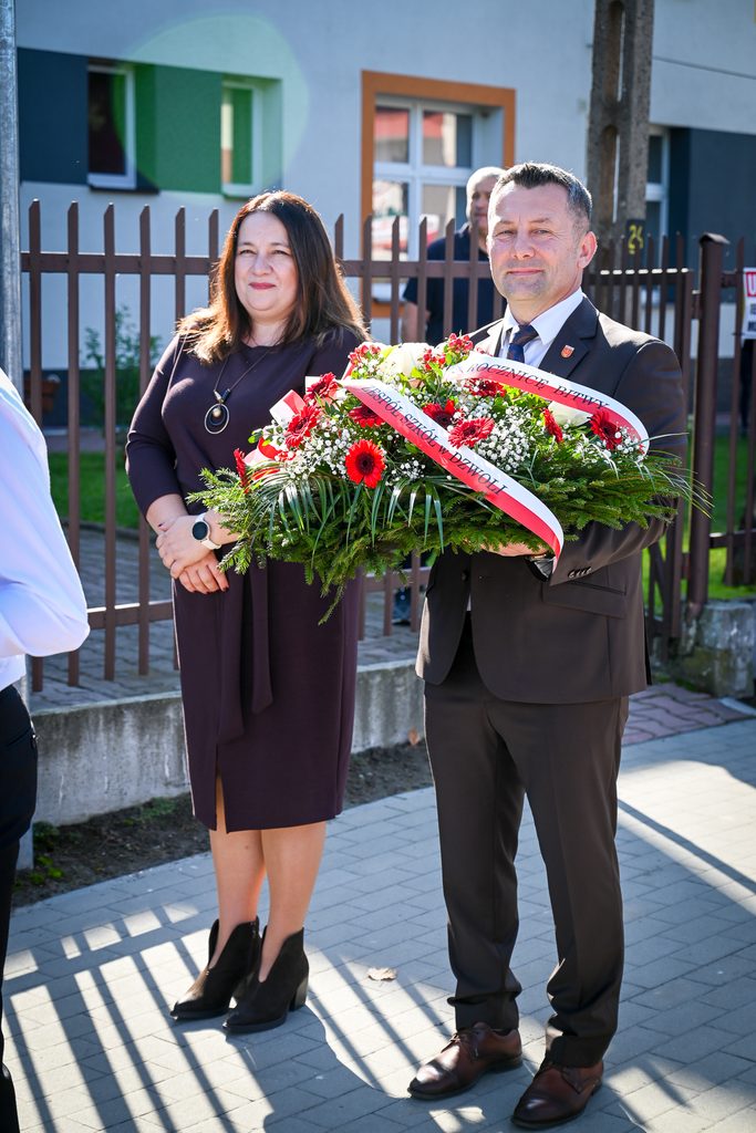 Mężczyzna w ciemnym garniturze trzyma bukiet czerwonych i białych kwiatów z biało-czerwoną wstążką, obok stoi kobieta w fioletowej sukience i czarnych botkach.