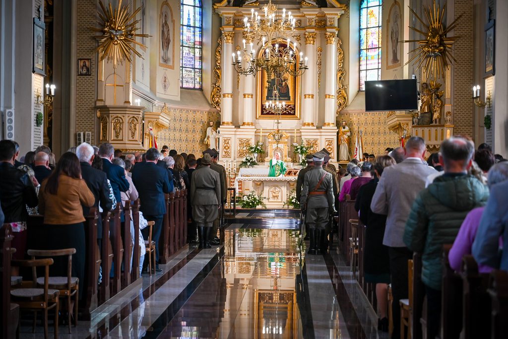 Wnętrze jasnej, zdobionej złotem świątyni z ludźmi stojącymi w ławkach, kapłanem w zielonych szatach przy ołtarzu i żołnierzami w mundurach.