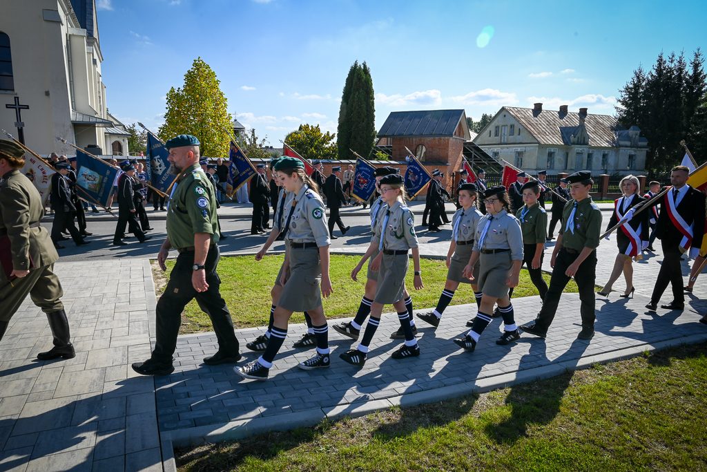 Grupa harcerzy i osób w mundurach maszerujących na tle budynków, niebieskie niebo, zielona trawa, flagi w tle.