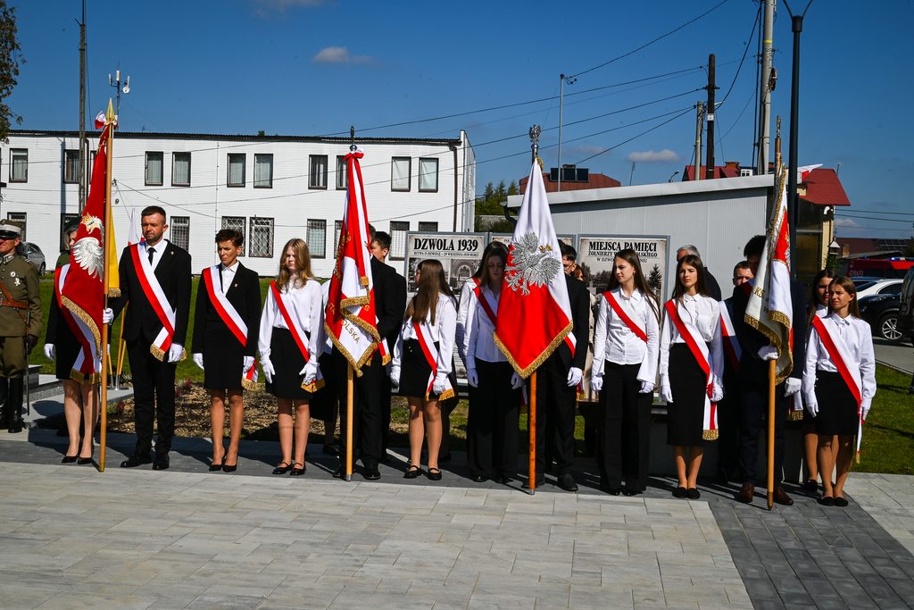 Grupa młodych ludzi w białych koszulach i czarnych spódnicach/spodniach, z biało-czerwonymi szarfami, trzymająca flagi z orłem na tle jasnego budynku i niebieskiego nieba.