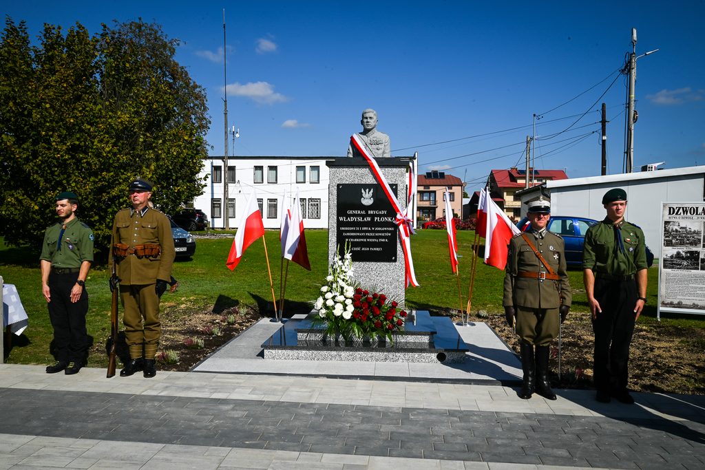 Pomnik generała Władysława Płonki z tablicą i kwiatami, cztery osoby w mundurach po bokach, biało-czerwone flagi, niebieskie niebo, budynki w tle.