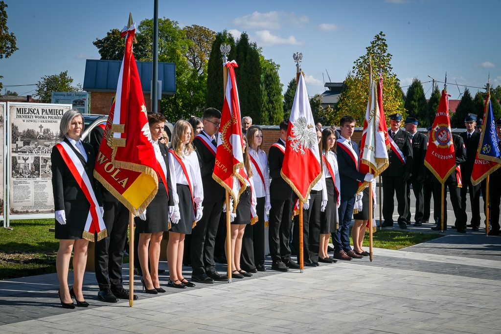 Grupa osób w mundurach i strojach formalnych trzymająca sztandary z orłami, na tle drzew i budynków, pod błękitnym niebem.