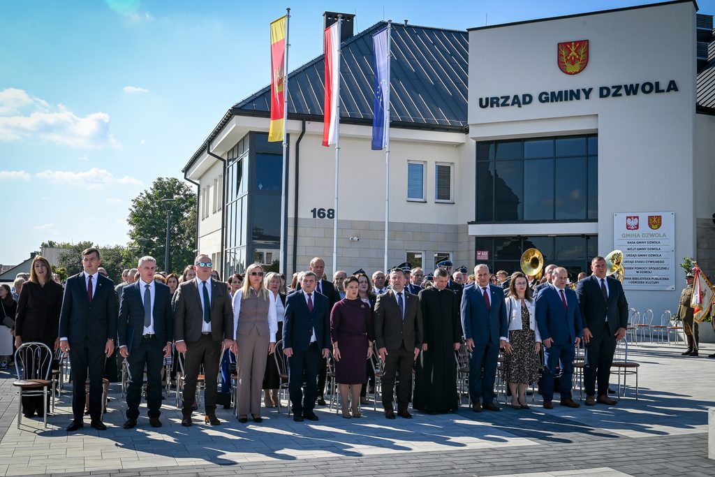 Grupa ludzi w formalnych strojach stoi przed budynkiem Urzędu Gminy Dzwola, za nimi trzy wysokie maszty z flagami, jasne niebo, szare płyty na placu.