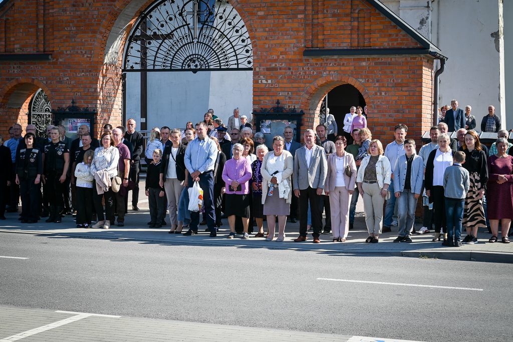 Grupa ludzi w różnym wieku stoi na chodniku przed budynkiem z cegły i dużymi łukowymi drzwiami z ozdobną kratą. Ubrani w ubrania codzienne i formalne.