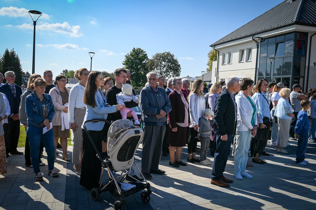 Grupa ludzi w różnym wieku stojąca na tle budynku i nieba, kobieta z wózkiem i dzieckiem, ubrani w jasne i ciemne ubrania, słoneczny dzień.