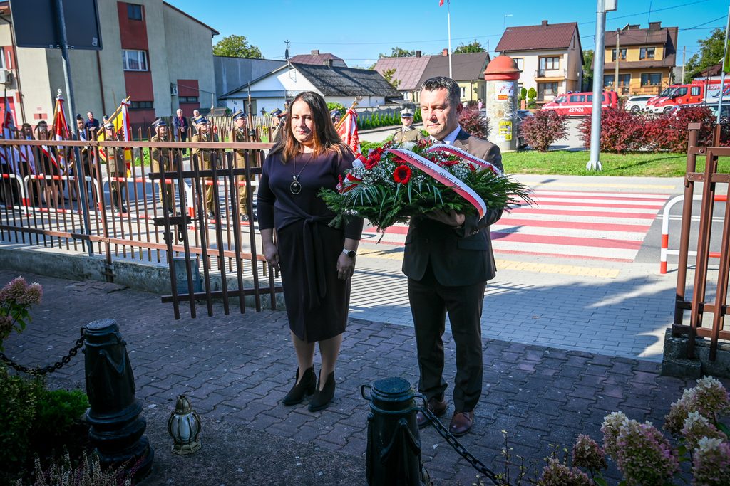 Mężczyzna w garniturze i kobieta w czarnej sukience stoją przed ogrodzeniem, mężczyzna trzyma bukiet czerwonych kwiatów z biało-czerwoną wstążką, w tle grupa osób w mundurach i flagi.