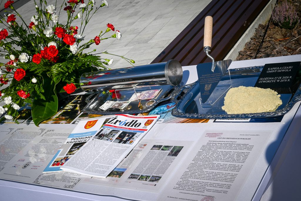 Bukiet czerwonych i białych goździków, metalowa kapsuła czasu, kielnia z drewnianą rączką, stos dokumentów i gazet na białym stole.