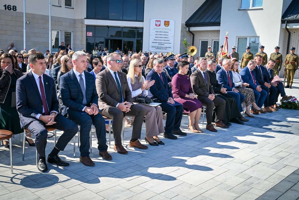 Grupa siedzących osób w formalnych strojach na zewnątrz przed budynkiem z napisem "Gmina Dzwola", w tle mundurowi i orkiestra dęta.
