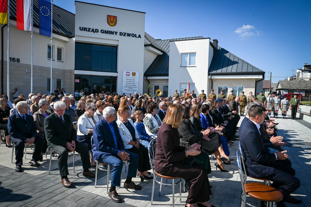 Ludzie siedzący na krzesłach przed budynkiem Urzędu Gminy Dzwola, po prawej stronie stoją żołnierze i harcerze w mundurach, niebieskie niebo.