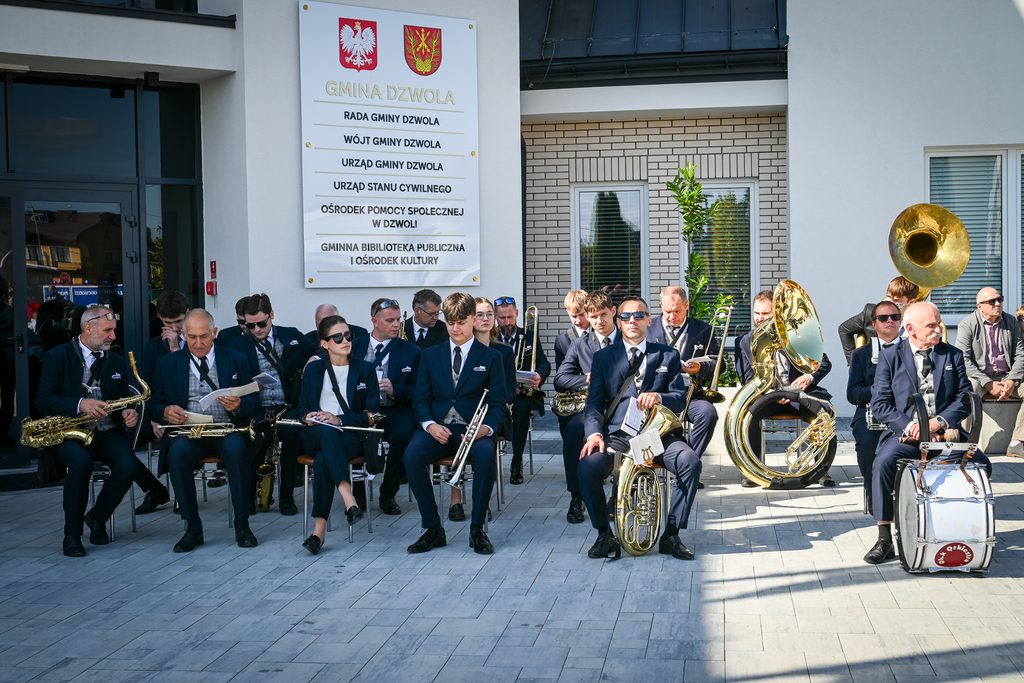 Grupa muzyków w granatowych garniturach siedzi z instrumentami dętymi i perkusyjnymi na tle białego budynku z tablicą informacyjną.