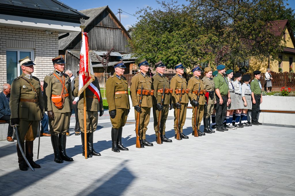 Grupa ludzi w mundurach historycznych i harcerskich stojąca w szeregu na tle budynków i zielonych drzew pod jasnym niebem.