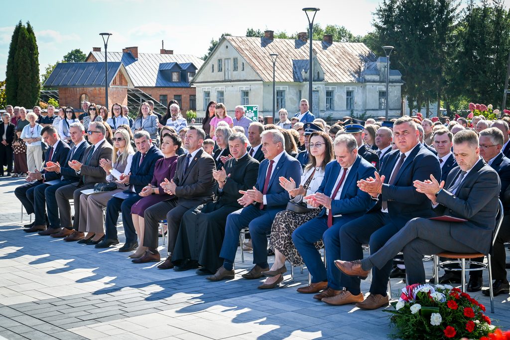 Grupa ludzi siedzących na krzesłach na zewnątrz, ubrani formalnie, w tle stare budynki i stojące osoby, jasne niebo, kwiaty na pierwszym planie.