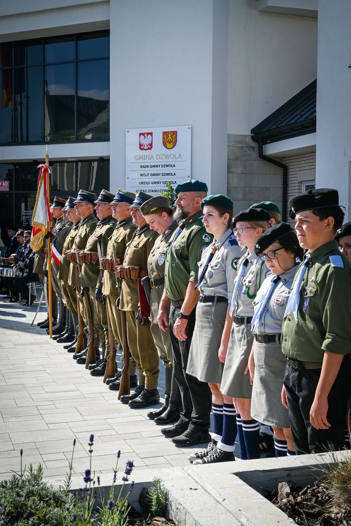 Linia osób w mundurach wojskowych i harcerskich, stojących na tle jasnego budynku z napisem "Gmina Dzwola", z flagą i bronią.