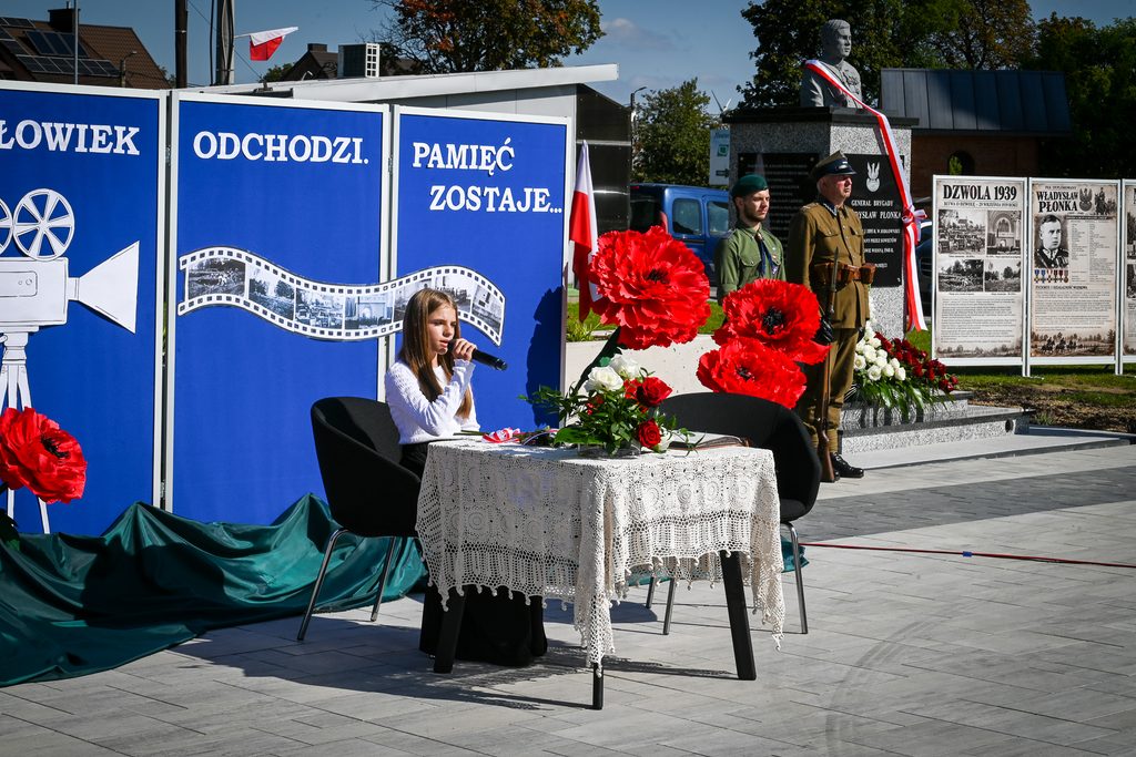 Dziewczynka z mikrofonem siedzi przy stoliku z białym koronkowym obrusem, za nią niebieskie plansze z napisem, obok pomnik, dwaj żołnierze i duże czerwone kwiaty.