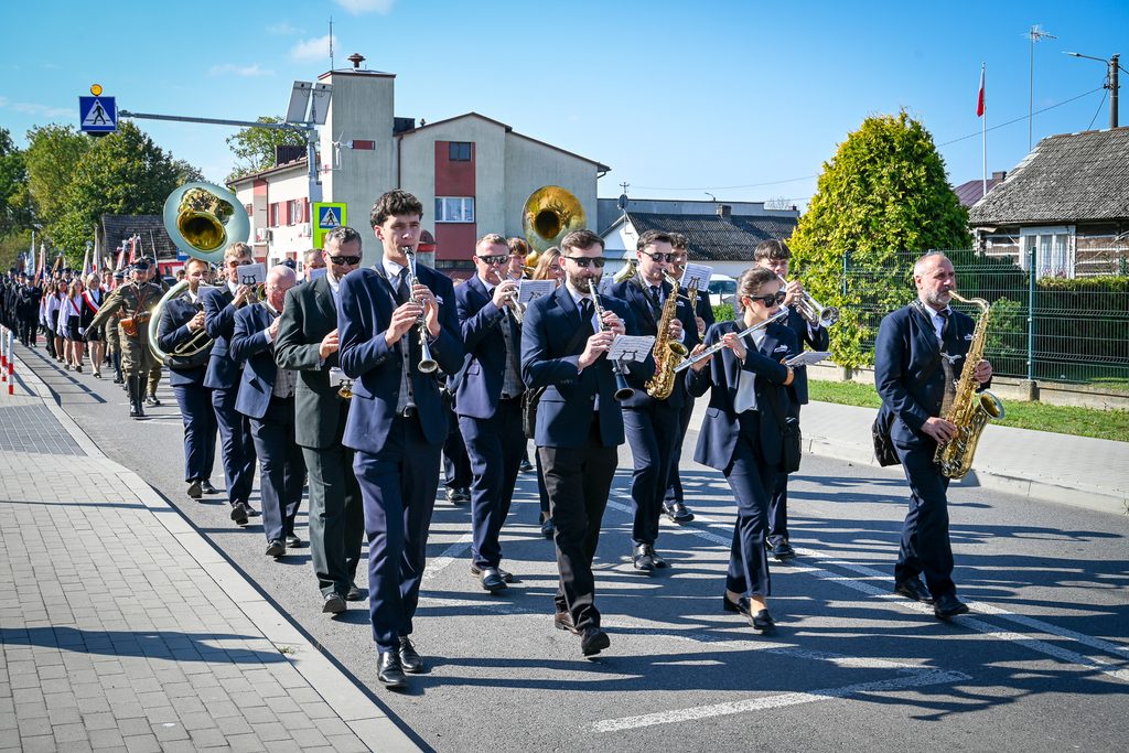 Marszujący muzycy w granatowych garniturach grający na instrumentach dętych, w tle ludzie z flagami i budynki mieszkalne pod błękitnym niebem.