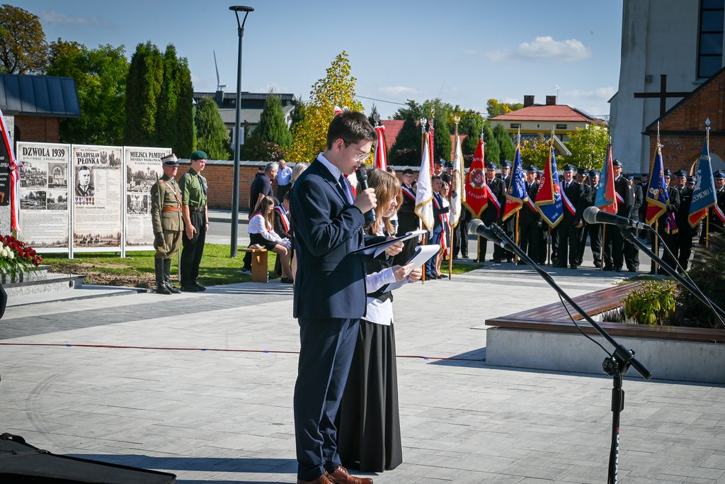 Młody mężczyzna i kobieta w eleganckich strojach czytają z kartki przy mikrofonie na zewnątrz, w tle ludzie w mundurach i flagi, tablice informacyjne i drzewa.
