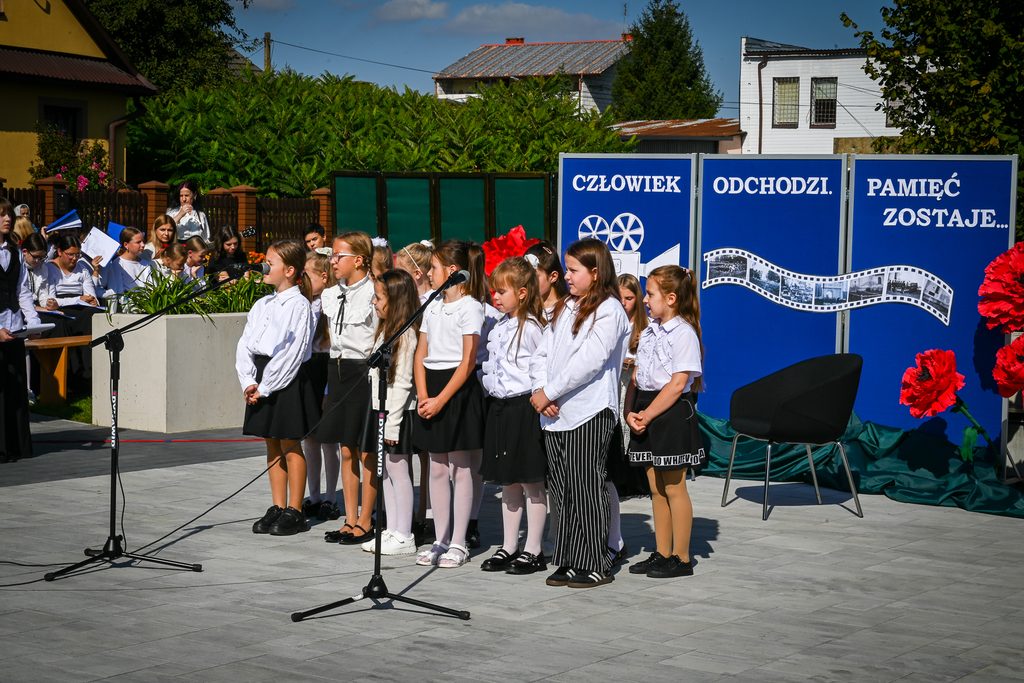 Grupa dziewczynek w białych bluzkach i czarnych spódnicach stoi przed niebieskimi planszami z białym napisem, na tle zielonych krzewów i budynków, mikrofony, czerwone dekoracje kwiatowe.