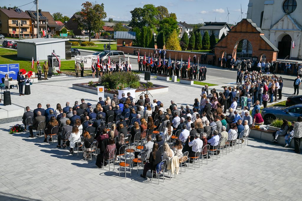 Ludzie siedzący na metalowych krzesłach na placu z jasną nawierzchnią, w tle grupy osób z flagami i pomnik, za nimi budynki mieszkalne i kościół z czerwonym ceglastym murem.