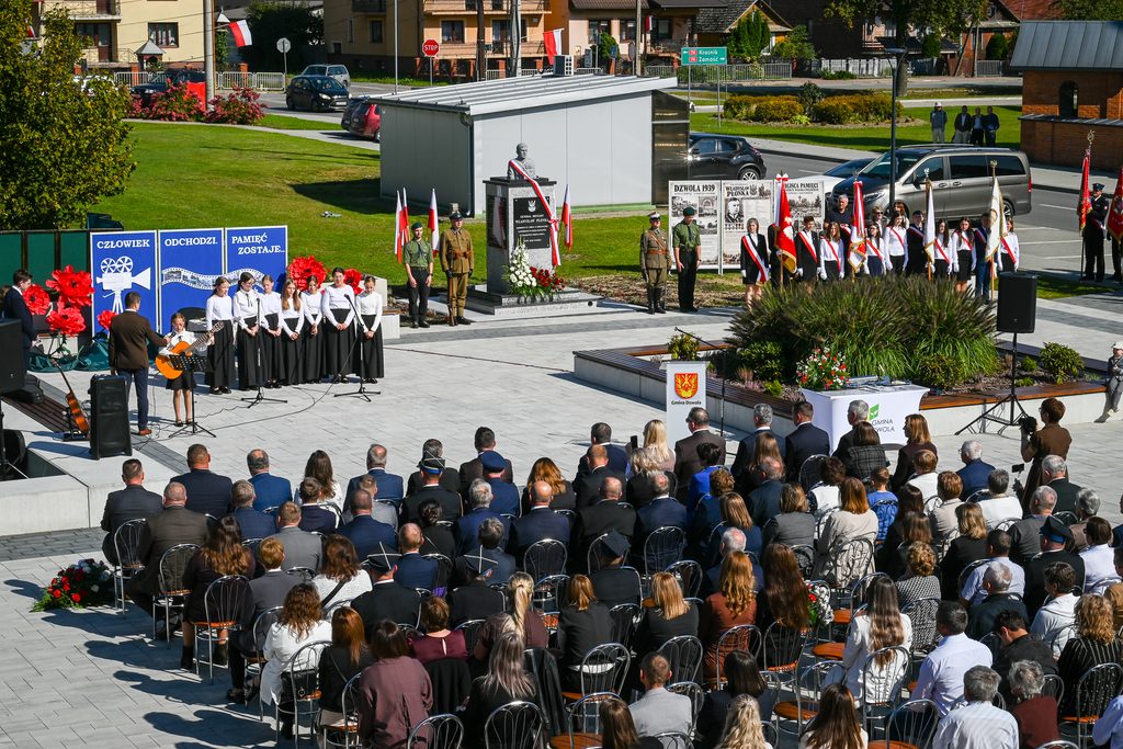 Uroczystość przy pomniku z biało-czerwonymi flagami, grupy ludzi w mundurach i białych koszulach, publiczność siedząca na krzesłach na placu z szarymi płytami.