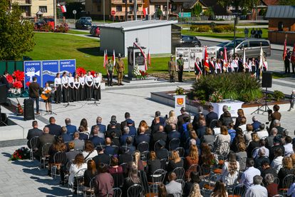 Uroczystość przy pomniku z biało-czerwonymi flagami, grupy ludzi w mundurach i białych koszulach, publiczność siedząca na krzesłach na placu z szarymi płytami.