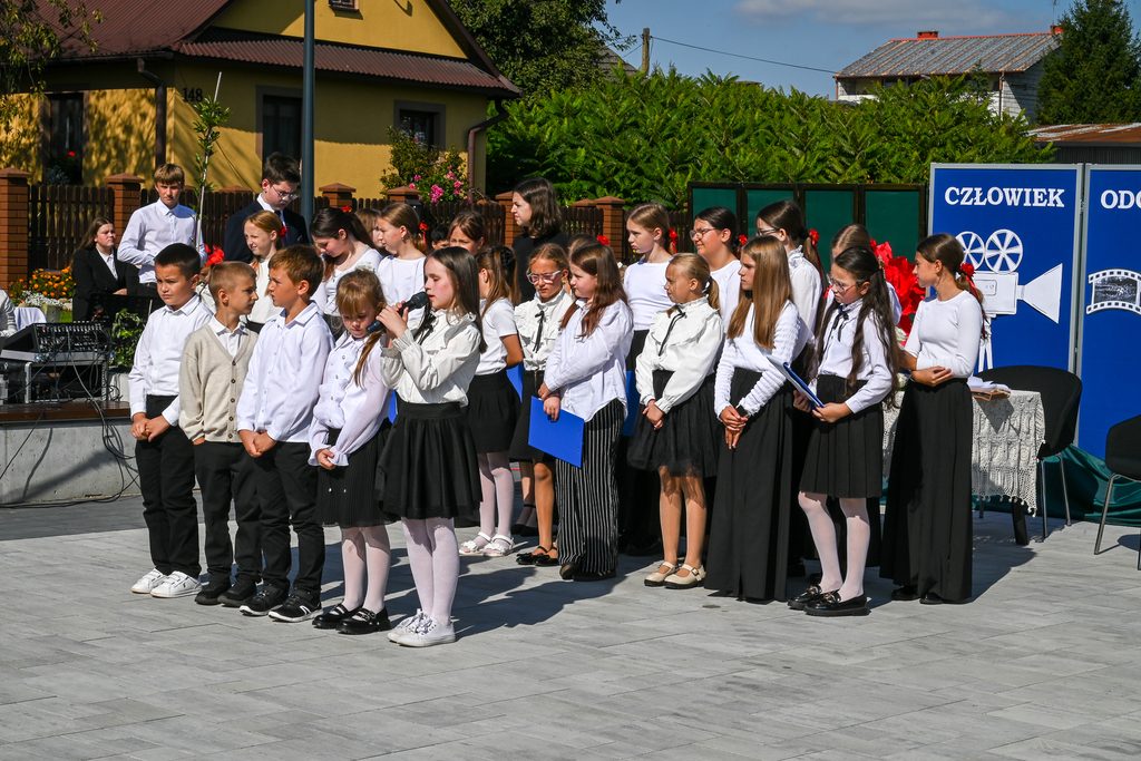 Grupa dzieci w białych bluzkach i ciemnych spodniach lub spódnicach stoi na zewnątrz na szarej, brukowanej nawierzchni, jedna dziewczynka mówi do mikrofonu.