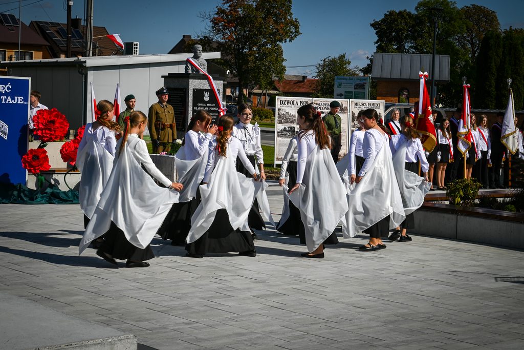 Grupa dziewcząt tańczących na placu, ubrane w białe bluzki i czarne spódnice z białymi pelerynami, w tle pomnik z biało-czerwoną szarfą i stojący ludzie z flagami.